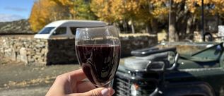 Hand hält ein Glas Rotwein vor einem offenen Geländewagen und leuchtend gelben Herbstbäumen.