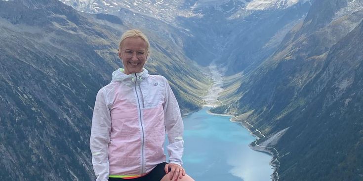 Simone Lorenz steht auf einem Bergplateau mit Blick auf einen türkisfarbenen Bergsee und schneebedeckte Gipfel in Norwegen