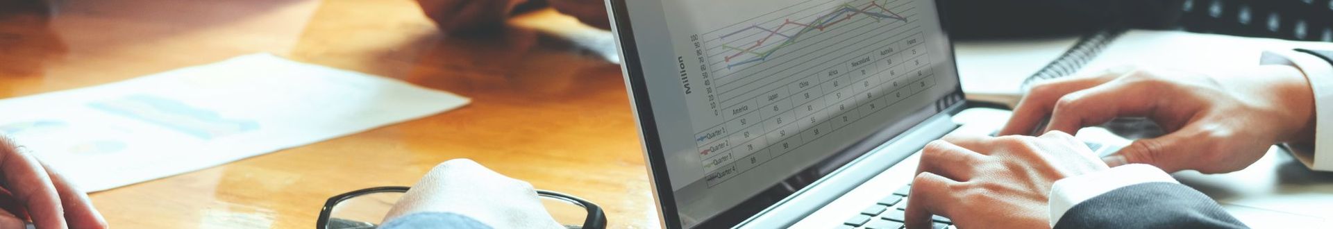 Mehrere Personen sitzen an einem Konferenztisch und arbeiten mit Laptop, Tablet und ausgedruckten Diagrammen. Auf dem Laptop-Display sind Finanzdiagramme und Tabellen zu sehen. Hände tippen auf der Tastatur, während andere Teilnehmende Unterlagen betrachten.