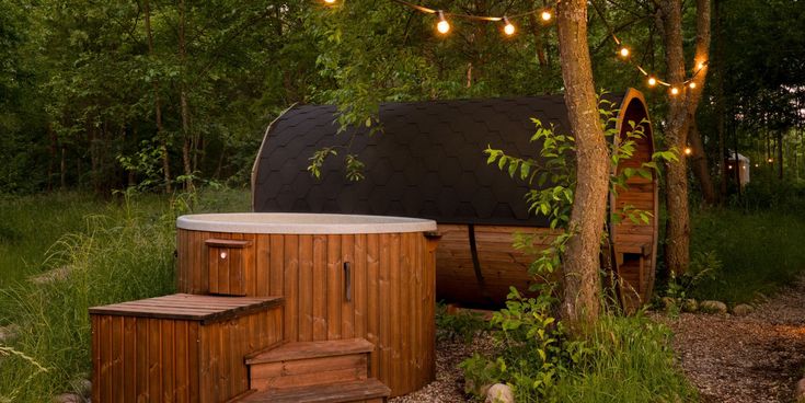 Ein runder Whirlpool aus Holz mit Einstiegstreppe steht neben einer fassförmigen Sauna im Wald, beleuchtet von warmen Lichterketten in den Bäumen.