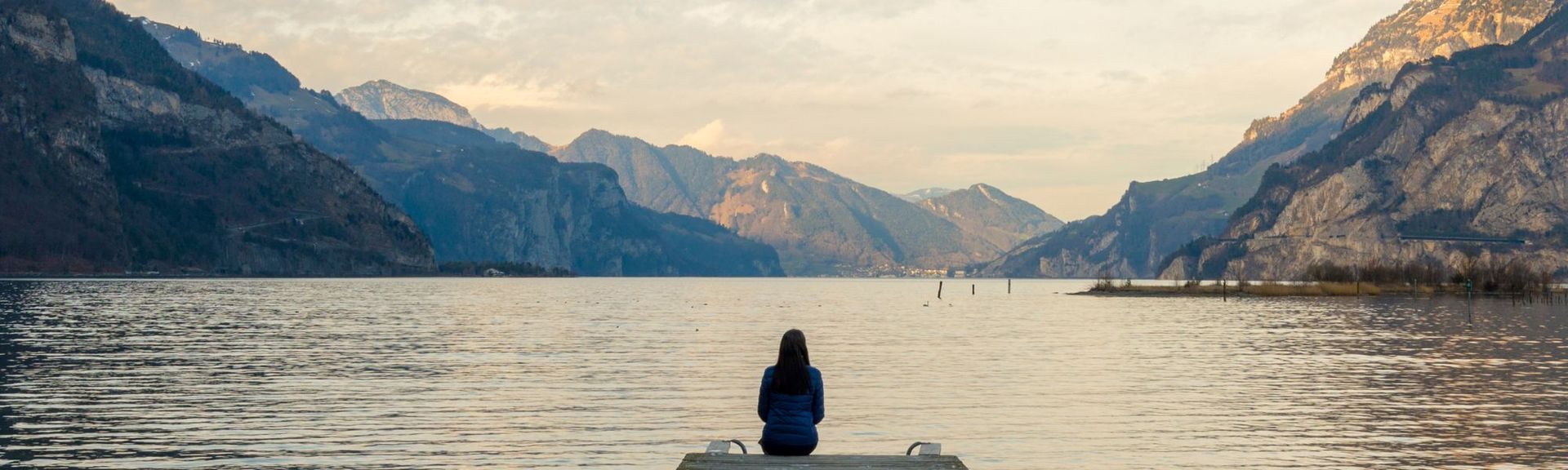 Person sitzt am Ende eines Stegs und blickt über einen ruhigen See, umgeben von Bergen – Sinnbild für Ruhe, Weitblick und Inspiration.