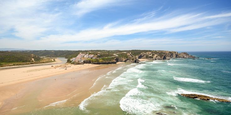 Weitläufiger Sandstrand an der Costa Vicentina mit türkisblauem Atlantik, felsiger Küste und kleiner Siedlung im Hintergrund.