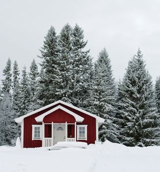 Ein kleines rotes Holzhaus steht umgeben von schneebedeckten Tannen in einer ruhigen, winterlichen Landschaft.