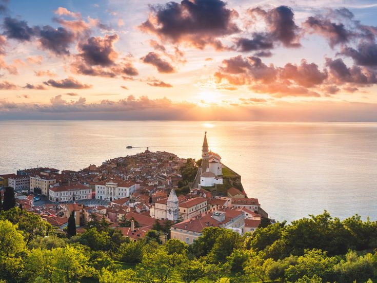 Ein malerischer Blick bei Sonnenuntergang auf die Küstenstadt Piran in Slowenien mit roten Dächern, einer Kirche auf der Landzunge und dem ruhigen Meer unter einem farbenprächtigen Himmel.