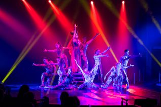Eine Gruppe von Artisten performt eine dynamische Bühnenshow unter farbigem Scheinwerferlicht in Rot, Pink und Gelb.