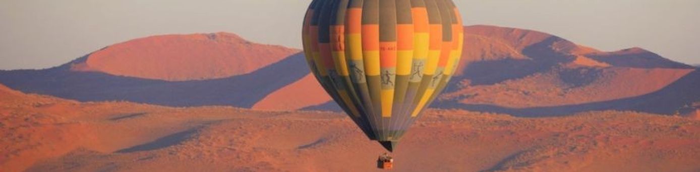 Heißluftballon schwebt über den roten Dünen einer Wüstenlandschaft bei Sonnenaufgang.
