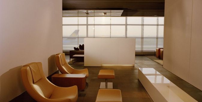 Lufthansa First Class Lounge mit ockerfarbenen Lounge-Sesseln und passenden Hockern, dunklem Steinboden und großer Fensterfront mit Blick aufs Rollfeld.