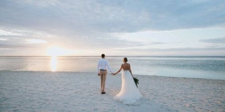 Ein Paar in Hochzeitskleidung spaziert Hand in Hand am Strand in den Sonnenuntergang – romantische Atmosphäre für Flitterwochen oder Strandhochzeiten.