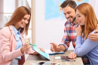 Beraterin erklärt einem jungen Paar Reiseunterlagen am Tisch – Beratungsgespräch in freundlicher Atmosphäre.