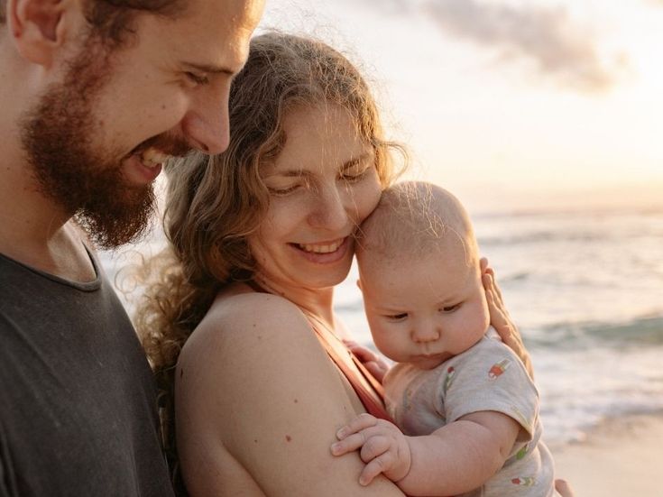 Junges Elternpaar hält liebevoll ihr Baby am Strand im Abendlicht – inniger Moment voller Geborgenheit und Glück.
