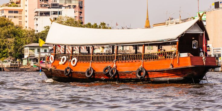 Traditionelles Holzboot mit weißem Sonnendach fährt auf einem Fluss in Bangkok, im Hintergrund moderne Gebäude und ein goldener Tempelturm.