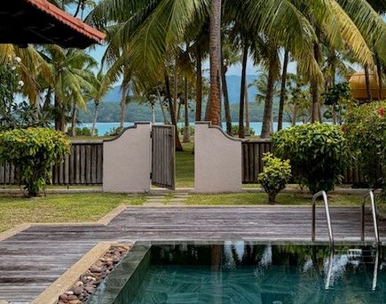 Poolanlage eines tropischen Resorts mit Palmen und Blick durch ein Gartentor auf das Meer.