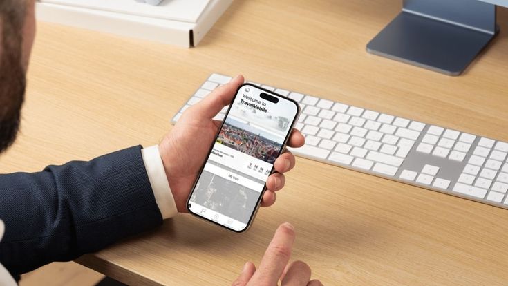 Geschäftsmann nutzt am Schreibtisch ein Smartphone mit geöffneter TravelMobile-App; im Hintergrund stehen Tastatur und Computerbildschirm.