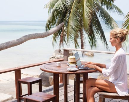 Frau sitzt auf Terrasse am Palmenstrand und arbeitet am Laptop