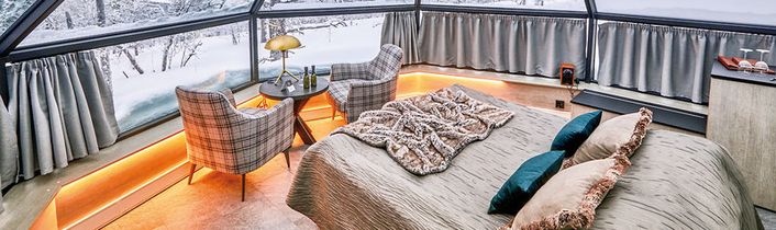 Gemütliches Schlafzimmer in einem Glasiglu mit Panoramablick auf eine verschneite Winterlandschaft bei Tageslicht