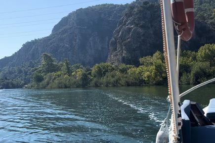 Blick von einem Boot auf ruhiges Wasser, bewaldete Felsen und blauen Himmel, Teil des Boots mit Rettungsring sichtbar.