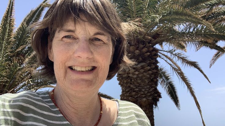 Petra Meyers lächelt vor einer großen Palme und dem blauen Meer auf Fuerteventura.