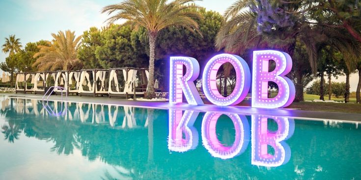 Beleuchtete Buchstaben am Pool des ROBINSON Clubs spiegeln sich im Wasser, umgeben von Palmen und balinesischen Daybeds – Symbol für stilvolle Entspannung und Urlaubsfeeling.
