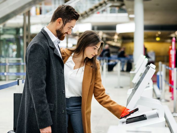 Mann und Frau am Self-Check-in-Automaten im Flughafenterminal; die Frau hält ihr Smartphone an den Scanner