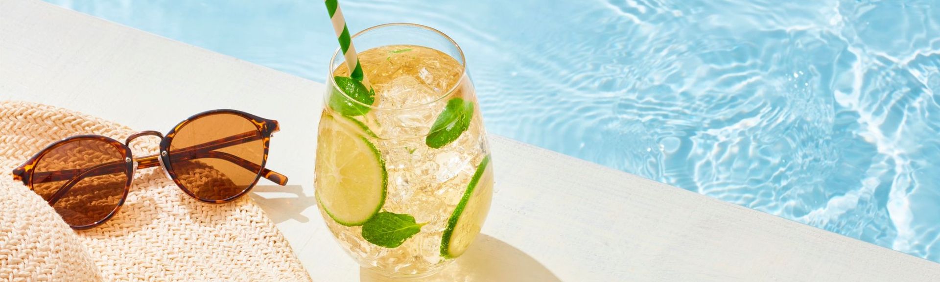 Sommerliche Szene am Pool mit Strohhut, Sonnenbrille und erfrischendem Getränk mit Limette und Minze auf einem hellen Tisch.