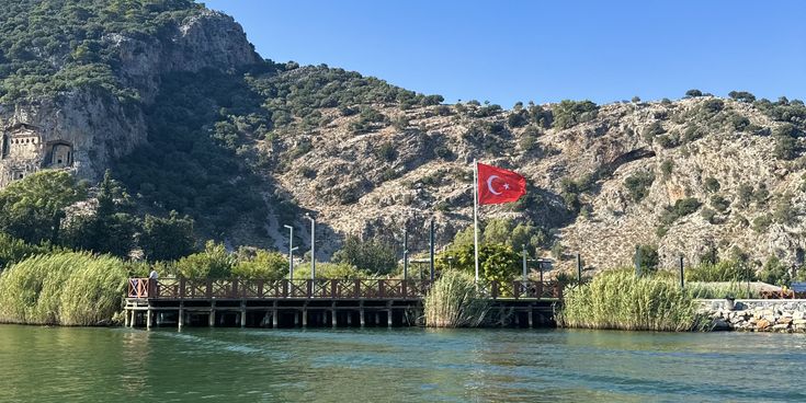 Türkische Flagge auf Fahnenmast am Ufer mit bewaldetem Hügel im Hintergrund und grünem Wasser im Vordergrund