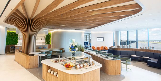 SkyTeam Flagship Lounge Frankfurt mit geschwungener Holzdecke, zentralem Buffet, Sitzbereichen und Fensterblick aufs Rollfeld.
