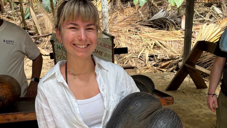 Georgina Dörling hält lächelnd eine große Coco-de-Mer-Nuss in einem tropischen Garten auf den Seychellen.