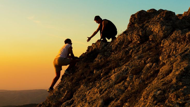 Zwei Personen beim Klettern auf einem Felsen im warmen Licht des Sonnenuntergangs – Symbol für Motivation, Zusammenhalt und gegenseitige Unterstützung auf dem Weg nach oben.