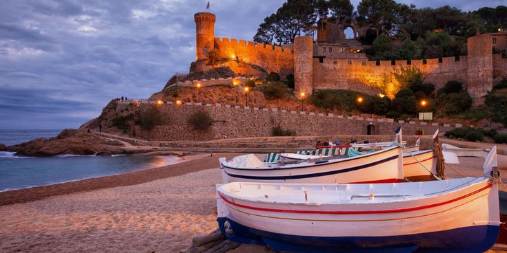Abendstimmung am Strand von Tossa de Mar an der Costa Brava, mit beleuchteter Burgmauer und Fischerbooten im Vordergrund.