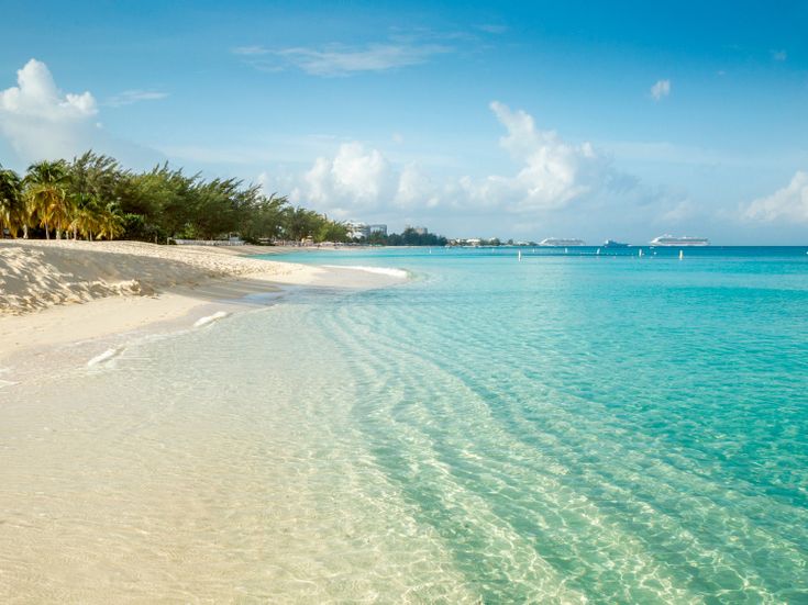 Weißer Sandstrand mit kristallklarem Wasser, im Hintergrund sind man vage ein paar Kreuzfahrtschiffe am Hafenanleger