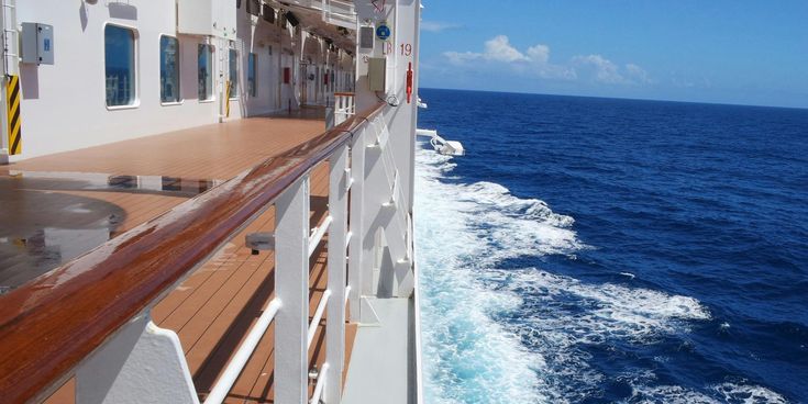 Seitliches Deck eines Kreuzfahrtschiffs mit Reling und Holzbelag, Blick auf tiefblaues Meer und weiße Bugwellen bei sonnigem Himmel.