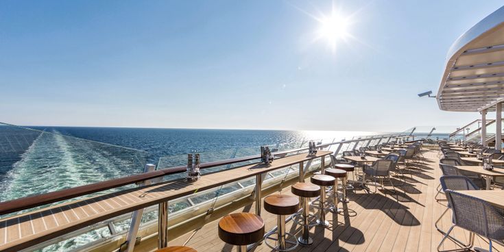 Sonnendeck eines Kreuzfahrtschiffs mit Barhockern und Tischen, Blick auf das Meer und die Heckwelle unter strahlender Sonne.