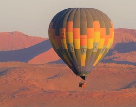 Heißluftballon schwebt über den roten Dünen einer Wüstenlandschaft bei Sonnenaufgang.