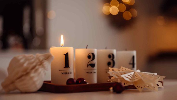 Eine brennende Adventskerze mit der Zahl eins steht auf einem Holztablett neben drei unangezündeten Kerzen und weihnachtlicher Dekoration in warmem Lichterglanz.