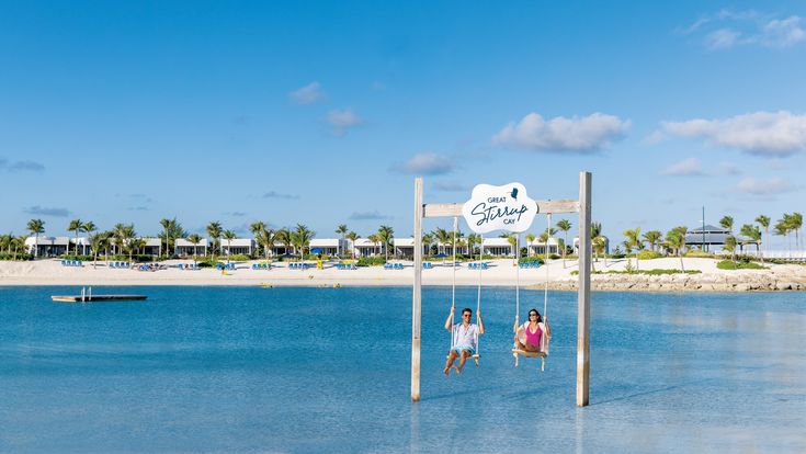 Zwei Personen schaukeln über dem türkisblauen Meer auf einer Holzschaukel mit dem Schild "Great Stirrup Cay", während im Hintergrund ein tropischer Sandstrand mit Palmen, Sonnenliegen und modernen Strandvillen unter blauem Himmel zu sehen ist.