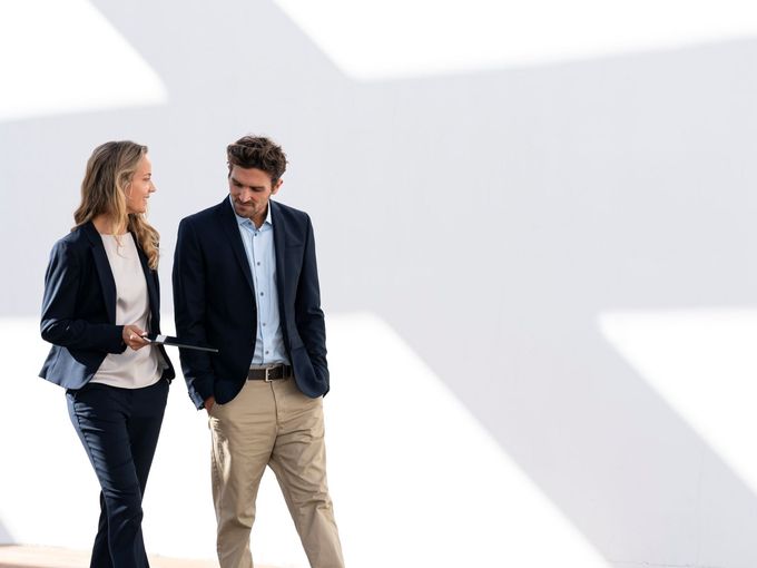 Zwei Geschäftsleute in Businesskleidung gehen nebeneinander und sprechen; die Frau hält ein Tablet in der Hand; vor hellem, minimalistischem Hintergrund.