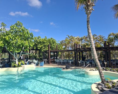Heller Hotelpool mit Palmen und angrenzendem Restaurantbereich unter blauem Himmel.