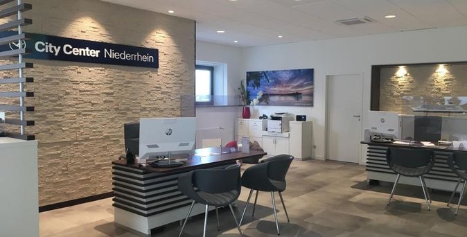 Moderner Arbeitsplatz in einem Reisebüro mit Holztisch, zwei Besucherstühlen und einem Modellkreuzfahrtschiff als Dekoration vor beleuchteter Steinwand.