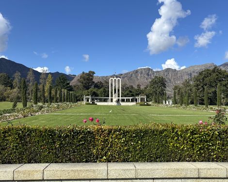 Weitläufiger Garten mit Rosenbeeten, zentralem Denkmal und den Bergen von Franschhoek im Hintergrund.