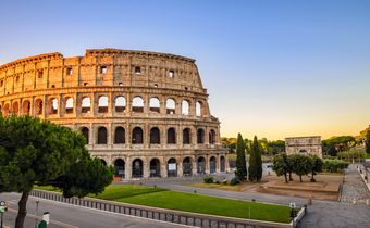 Kolosseum in Rom im warmen Abendlicht mit grünem Park und blauem Himmel im Hintergrund.