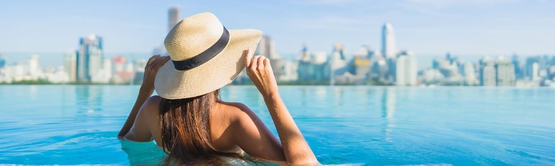Frau mit Strohhut steht in einem Infinitypool und blickt auf eine moderne Großstadtskyline.