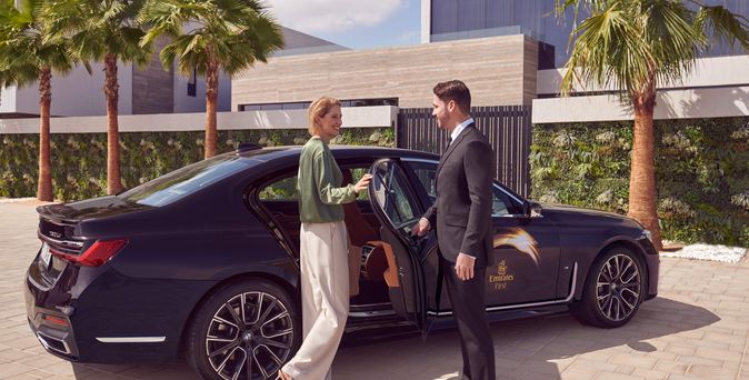 Ein Chauffeur im dunklen Anzug öffnet einer Frau die Autotür und begrüßt sie beim Einsteigen in eine schwarze Limousine.