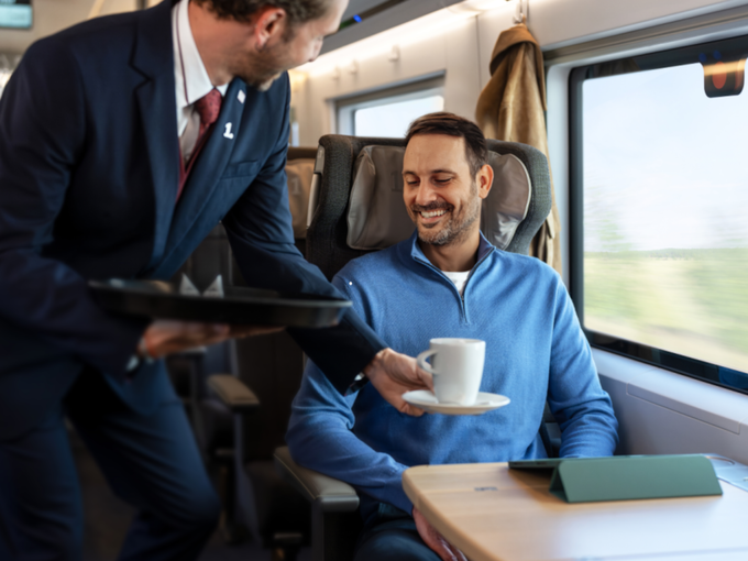 Zugbegleiter serviert einem lächelnden Fahrgast im 1.-Klasse-Abteil Kaffee, am Tisch liegt ein Tablet.