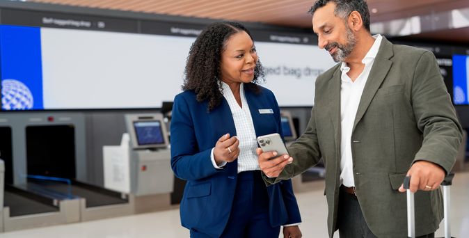 Eine Mitarbeiterin im blauen Blazer zeigt einem Reisenden mit Koffer etwas auf seinem Smartphone, im Hintergrund Self-Service-Check-in-Automaten.