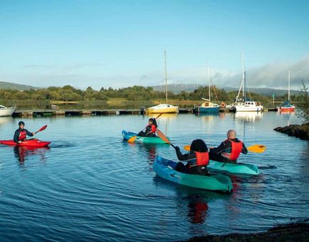Gruppe von Kajakfahrern paddelt auf einem klaren See umgeben von Bergen und Booten im Hintergrund.