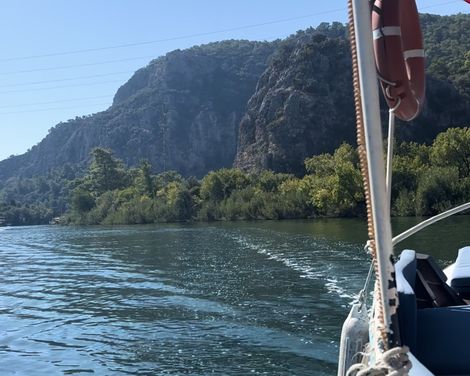 Blick von einem Boot auf ruhiges Wasser, bewaldete Felsen und blauen Himmel, Teil des Boots mit Rettungsring sichtbar.