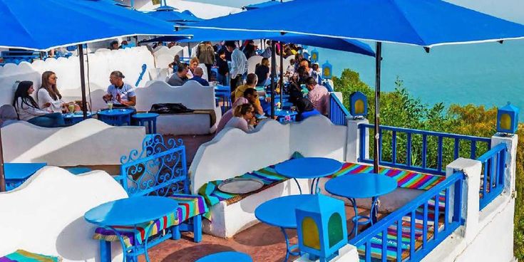 Menschen sitzen unter blauen Sonnenschirmen in einem farbenfrohen Café mit Blick auf das Meer – Symbol für mediterranes Flair und entspannte Urlaubsmomente in Nordafrika.