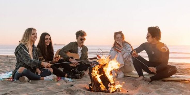 Gruppe junger Menschen sitzt am Strand um ein Lagerfeuer, lacht und spielt Musik – stimmungsvolles Motiv für gemeinsame Erlebnisse auf Reisen.