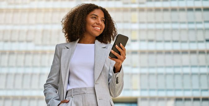Lächelnde Geschäftsfrau steht vor Bürogebäude mit Smartphone in der Hand