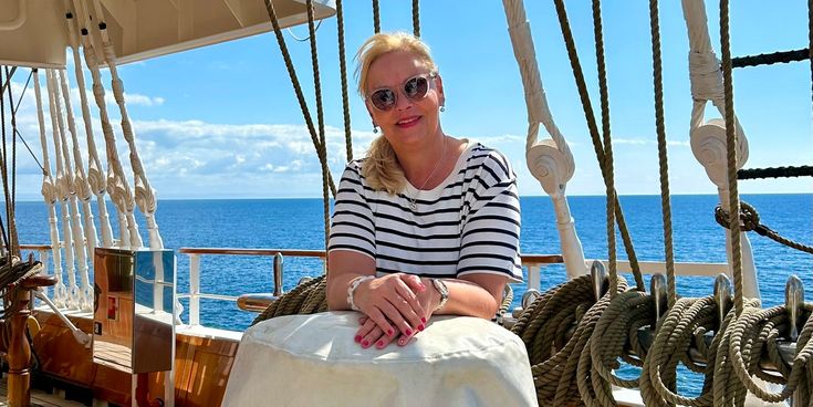 Christina sitzt entspannt an Deck eines Segelschiffs bei sonnigem Wetter mit Blick auf das offene Meer und Takelage im Hintergrund.